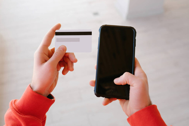 Nézőpont vágott közeli nézet fotó férfi kezét visel piros kapucnis üres hitelkártya és okostelefon. Online fizetési koncepció. - Fotó, kép
