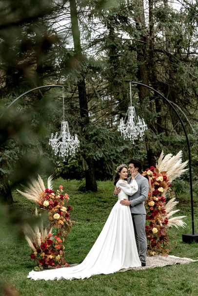 Красивая свадебная пара обнимается и целуется на свадебной церемонии на фоне леса. Высокое качество фото - Фото, изображение