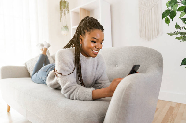 Young African American teen on a sofa in her living room using cellphone - Foto, afbeelding