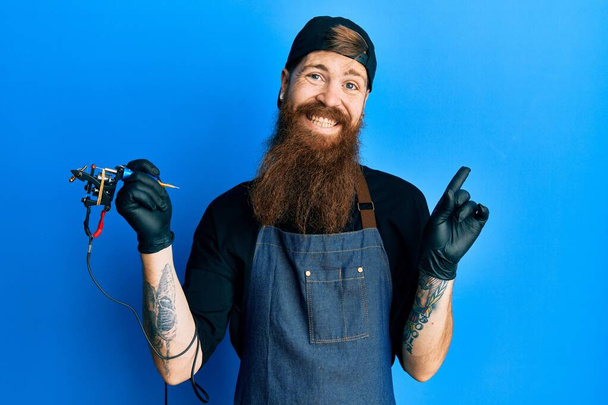 Redhead man with long beard tattoo artist wearing professional uniform and gloves cheerful with a smile on face pointing with hand and finger up to the side with happy and natural expression  - Foto, immagini
