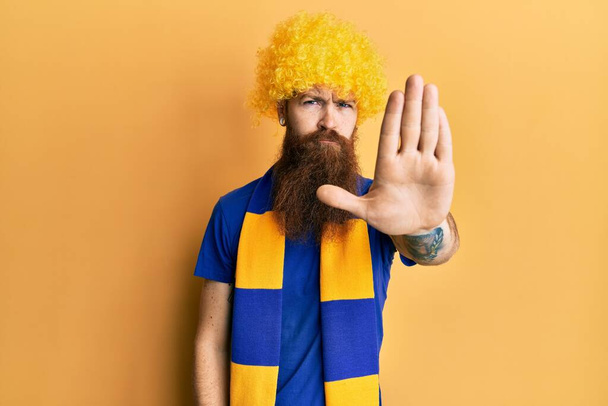 Roodharige man met lange baard voetbal hooligan juichen spel dragen grappige pruik doen stoppen met zingen met palm van de hand. waarschuwingsuitdrukking met negatief en ernstig gebaar op het gezicht.  - Foto, afbeelding