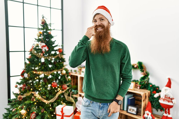 Redhead man with long beard wearing christmas hat by christmas tree pointing with hand finger to face and nose, smiling cheerful. beauty concept  - Foto, immagini
