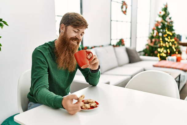 Young redhead man having breakfast sitting by christmas tree at home - Photo, Image