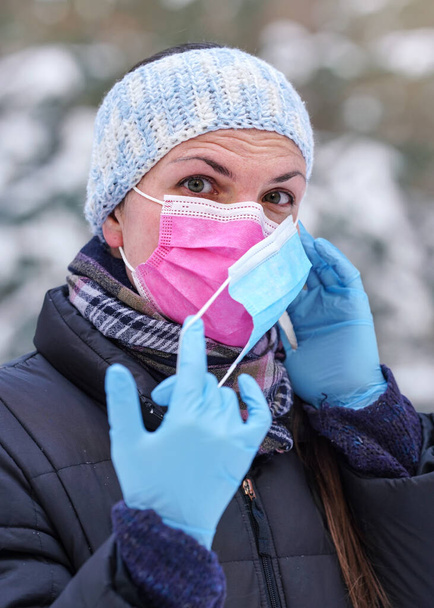 Young woman in warm winter clothing wearing pink disposable virus face mask, putting another one on - some advise that two layers provides better protection again coronavirus covid-19 spread - Zdjęcie, obraz