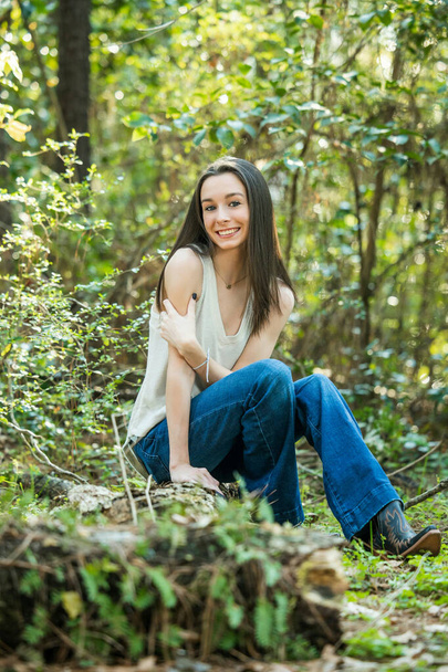 Une belle adolescente brune heureuse assise à l'extérieur sur une bûche hapy et souriante avec espace de copie - Photo, image