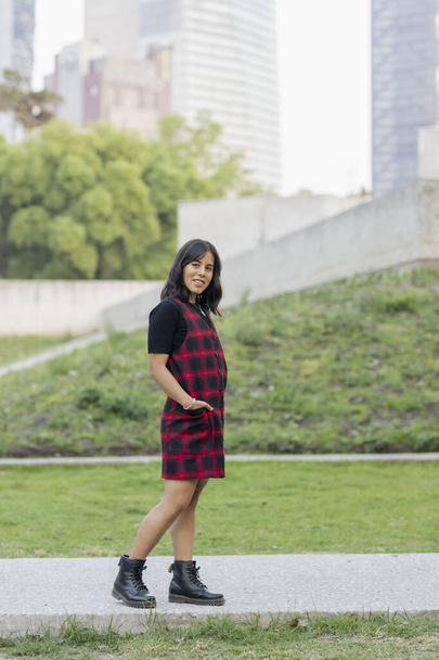 portrait of a young latin woman with a red dress, in a park with tall buildings in the background - Photo, Image