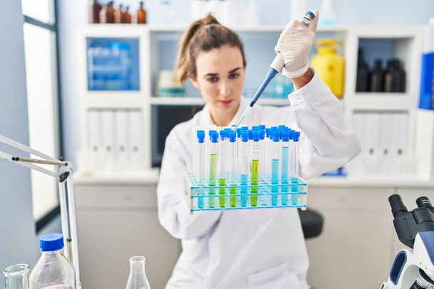 Jovem mulher vestindo uniforme de cientista usando pipeta trabalhando em laboratório - Foto, Imagem