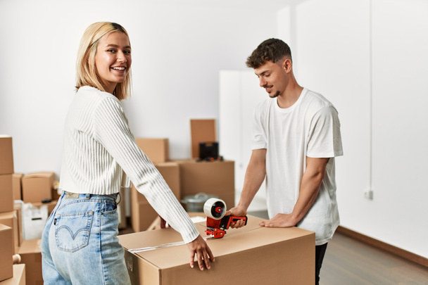 Молода біла пара посміхається щаслива закриваюча коробка з використанням пакувальної стрічки в новому будинку
. - Фото, зображення