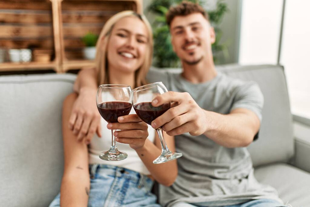 Joven pareja caucásica sonriendo feliz brindis con vino tinto en casa. - Foto, imagen