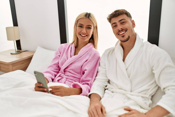 Joven pareja caucásica sonriendo feliz usando smartphone sentado en la cama en casa. - Foto, imagen
