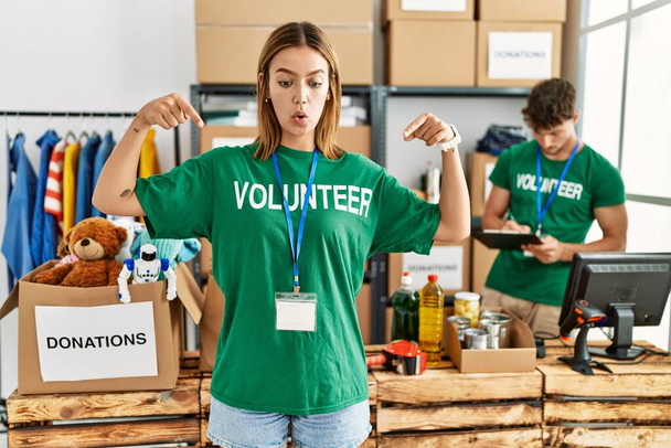 Junges blondes Mädchen im freiwilligen T-Shirt am Spendenstand zeigt mit nach unten zeigenden Fingern Werbung, überraschtem Gesicht und offenem Mund  - Foto, Bild