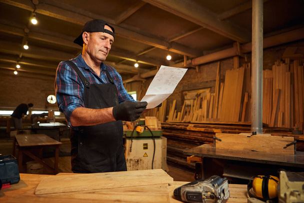 Πορτρέτο ενός παλαιού ξυλουργού που εξετάζει σχέδια για να κάνει ένα έπιπλο στο εργαστήριο ξυλουργικής - Φωτογραφία, εικόνα