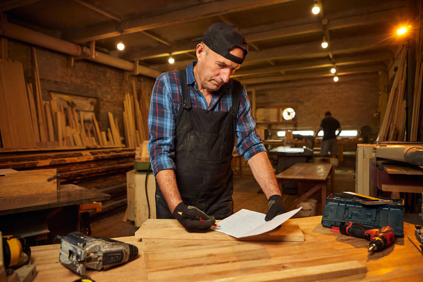 Портрет старшего плотника, рассматривающего чертежи, планирует изготовить мебель в столярной мастерской - Фото, изображение