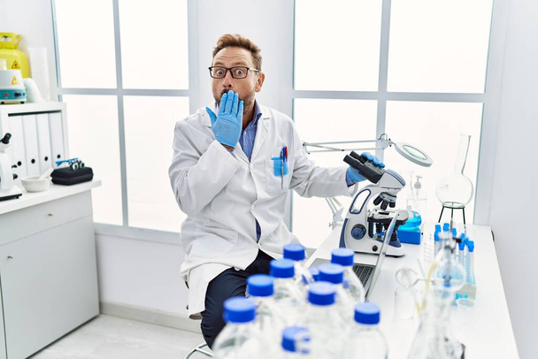 Hombre de mediana edad que trabaja en el laboratorio científico cubriendo la boca con la mano, sorprendido y temeroso de equivocarse. expresión sorprendida  - Foto, Imagen