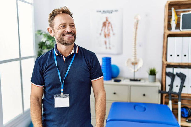 Middle age physiotherapist man working at pain recovery clinic looking away to side with smile on face, natural expression. laughing confident.  - Фото, изображение