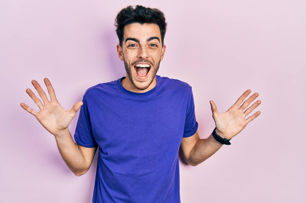Νεαρός Ισπανόφωνος φορώντας casual t-shirt γιορτάζει τρελός και έκπληκτος για την επιτυχία με τα χέρια σηκωμένα και ανοιχτά μάτια ουρλιάζοντας ενθουσιασμένος. έννοια νικητή  - Φωτογραφία, εικόνα