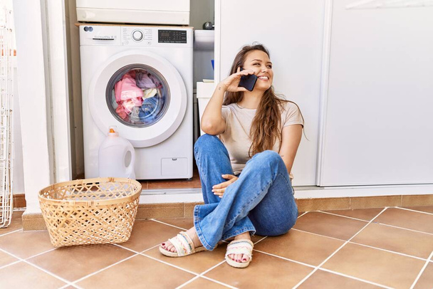 Mladý hispánec dívka mluví na smartphone čeká na pračku doma - Fotografie, Obrázek