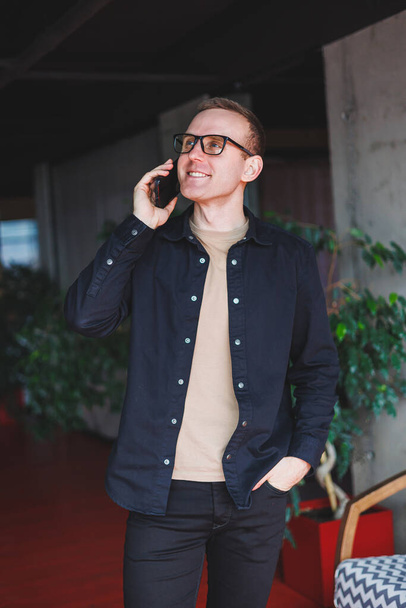 Портрет щасливого чоловіка в оптичних окулярах, що тримає сучасний мобільний телефон і посміхається на камеру, веселий блогер з цифровим смартфоном, який позує в коворкінгу
 - Фото, зображення