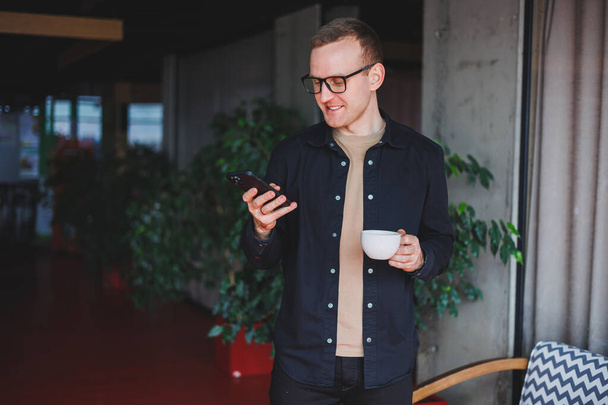 Portrait d'un heureux homme aux lunettes optiques tenant dans ses mains un téléphone portable moderne et un café, un blogueur joyeux avec un smartphone numérique posant dans un café - Photo, image