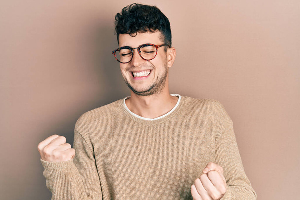 Junger hispanischer Mann in lässiger Kleidung und Brille sehr glücklich und aufgeregt dabei Siegergeste mit erhobenen Armen, lächelnd und schreiend nach Erfolg. Festkonzept.  - Foto, Bild