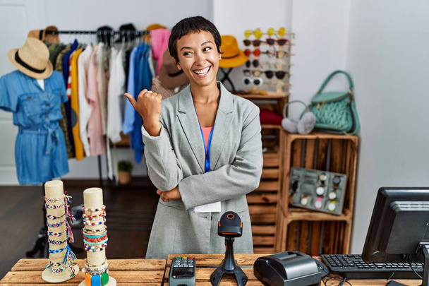 Young hispanic woman with short hair working as manager at retail boutique smiling with happy face looking and pointing to the side with thumb up.  - Photo, Image