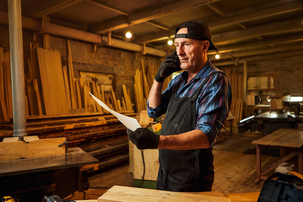 Portrait of a senior carpenter looking at blueprints plans and talking phone with a client in the carpentry workshop - Фото, изображение