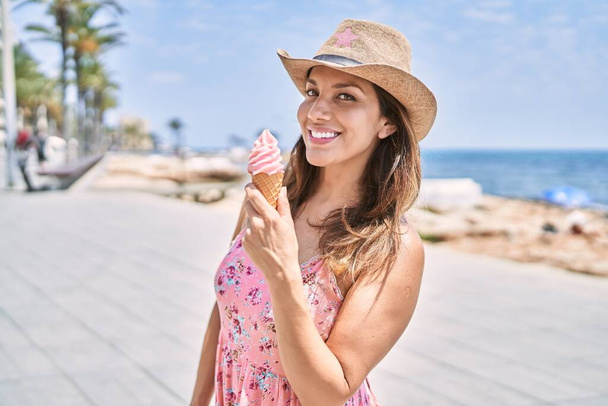 Μελαχρινή γυναίκα απολαμβάνει μια καλοκαιρινή μέρα στην παραλία τρώγοντας παγωτό χωνάκι. - Φωτογραφία, εικόνα