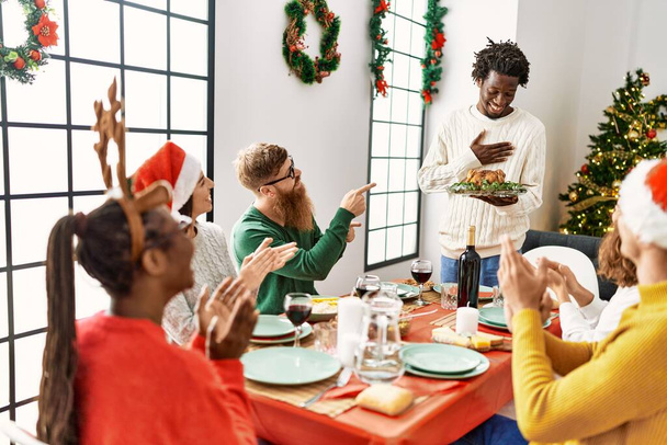 Ryhmä ihmisiä tapaamassa taputusta ja istumassa pöydällä. Mies seisoo ja pitää paahdettua kalkkunaa juhlimassa joulua kotona. - Valokuva, kuva