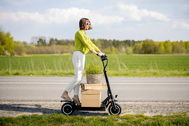 Woman driving electric scooter with parcels on the field road - Фото, изображение
