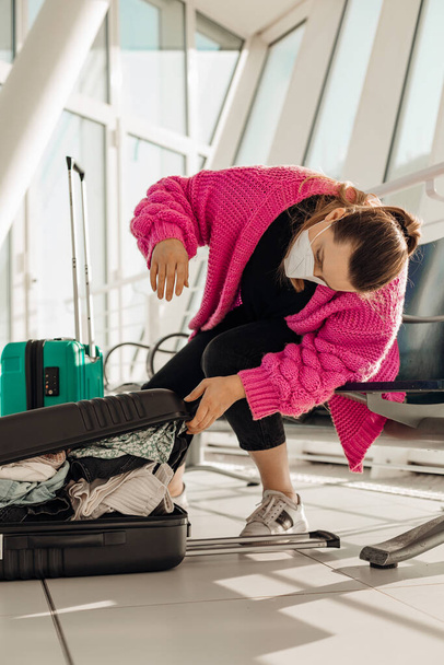 Függőleges nő rózsaszín dzsekiben, maszkot visel, csomagolja a poggyászát a reptéren a váróteremben. Késő a repüléshez. - Fotó, kép