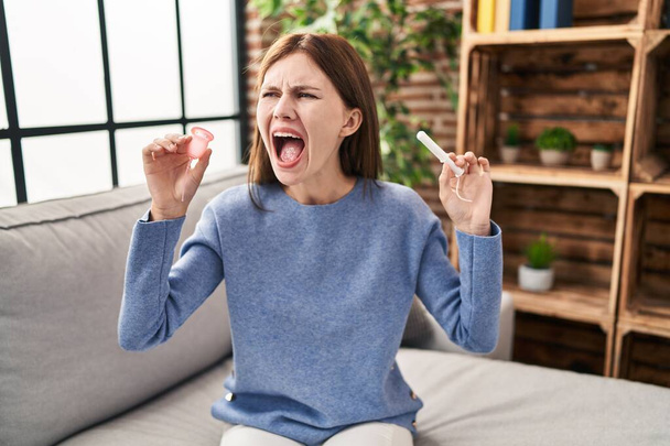 Jeune femme brune tenant la coupe menstruelle et tampon en colère et fou hurlant frustré et furieux, criant de colère. rage et concept agressif.  - Photo, image