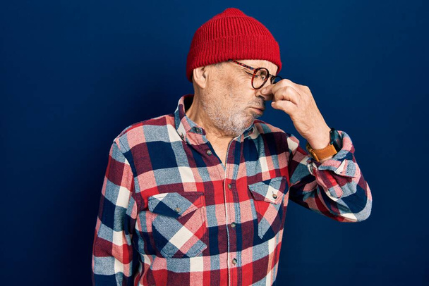 Handsome mature man wearing hipster look with wool cap smelling something stinky and disgusting, intolerable smell, holding breath with fingers on nose. bad smell  - Foto, afbeelding