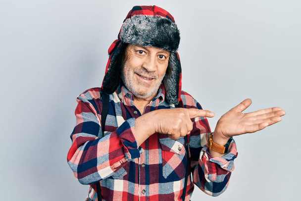 Bonito homem maduro usando chapéu de inverno com abas de orelha espantado e sorrindo para a câmera enquanto se apresenta com a mão e apontando com o dedo.  - Foto, Imagem