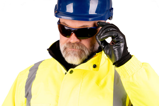 Een bouwvakker met een blauwe hoed en een gele hi-viz jas op een witte achtergrond met een donker getinte veiligheidsbril - Foto, afbeelding
