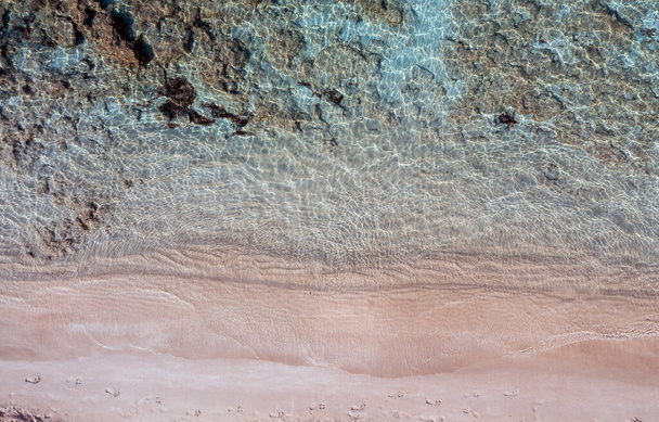 Homokos strand, türkiz kék színű tengeri antenna felülnézet. Lábnyomok nedves rózsaszín homokon. Tiszta, átlátszó víz, sziklás tengerfenék. Égei-tengeri görög sziget nyári vakáció destinatio - Fotó, kép