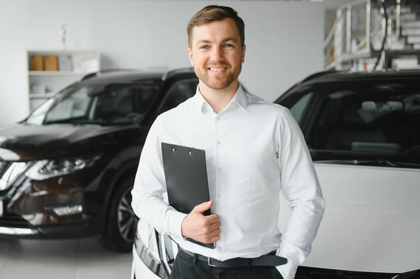 portrait of open-minded professional salesman in cars showroom, caucasian man in white formal shirt stands next to luxurious car and looks at camera. - Fotó, kép