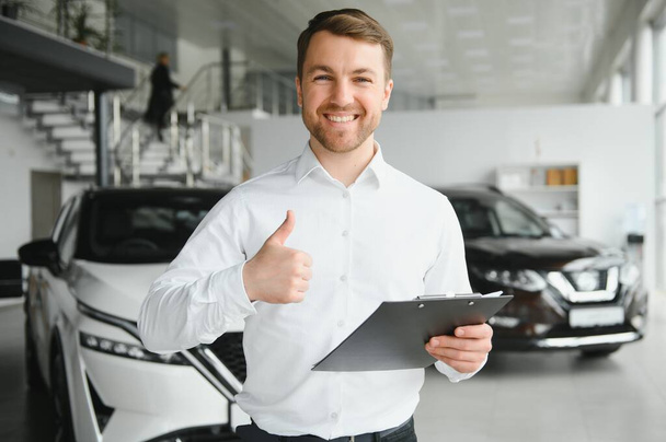 portrait of open-minded professional salesman in cars showroom, caucasian man in white formal shirt stands next to luxurious car and looks at camera. - Photo, image