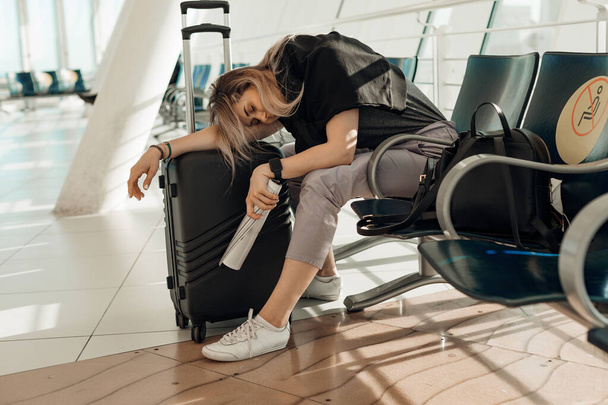 Tired and sleepy blond woman in casual clothes with luggage, leaning on bags, sitting in airport terminal. Cancel flight - Foto, Bild
