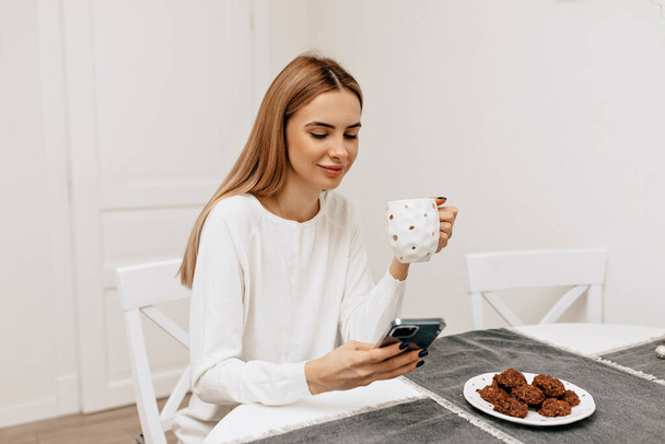 Charming pretty girl with long light hair is sitting in the kitchen and drinking tea. Lovable girl is having coffee break at home. High quality photo - Fotografie, Obrázek