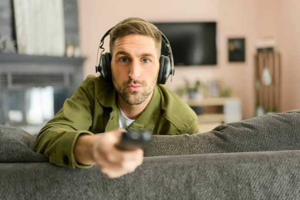 Όμορφος τύπος φορώντας ακουστικά απολαμβάνει τη μουσική στον άνετο καναπέ με τηλεχειριστήριο - Φωτογραφία, εικόνα