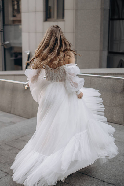 Vertical portrait of beautiful woman with long hair in white wedding dress on the city background. The bride stands with her back. Fashionable long sleeves white wedding dress with bare shoulders - Foto, Bild