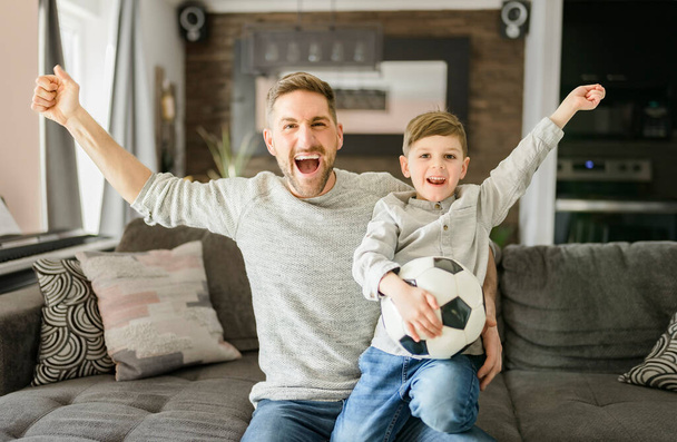 Soccer fans Emotional dad and son cheering with football ball, watching sport on tv at home - Φωτογραφία, εικόνα