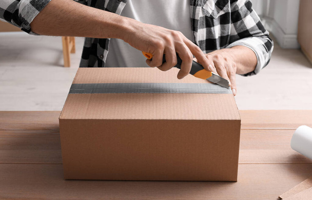 Man using utility knife to open parcel at wooden table, closeup - Photo, Image