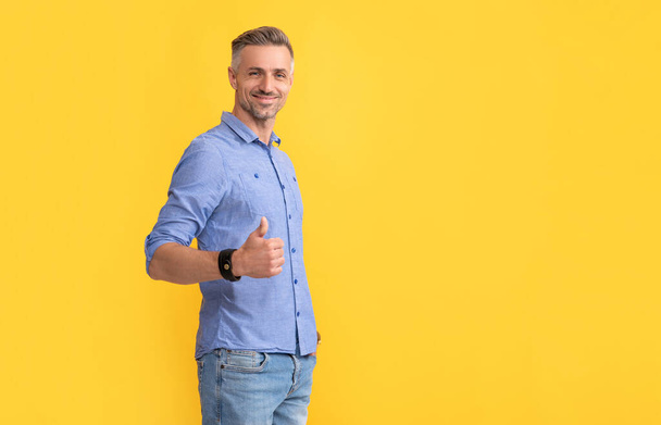 smiling mature man with grizzled hair show thumb up on yellow background, copy space, success - Photo, Image