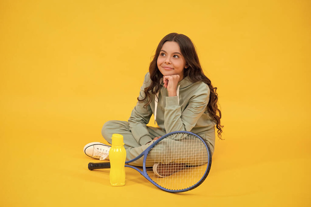 usměvavé dítě sedí ve sportovním oblečení s squash raketou a láhev vody na žlutém pozadí, sportovní - Fotografie, Obrázek