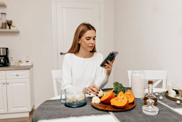 Bella donna attraente con i capelli scuri che indossano camicia bianca sta cercando nella ricetta smartphone. Donna in cucina con verdure fresche di cottura. Foto di alta qualità - Foto, immagini