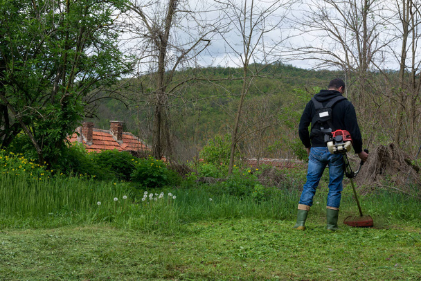 Een man maait het gras met een trimmer op een lentedag. Boer maait gras in een dorp in de achtertuin met handgrasmaaier - Foto, afbeelding