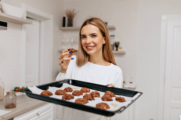 Charming smiling girl with happy smile holding baking tray with cookies and posing at camera at home kitchen. High quality photo - Fotografie, Obrázek