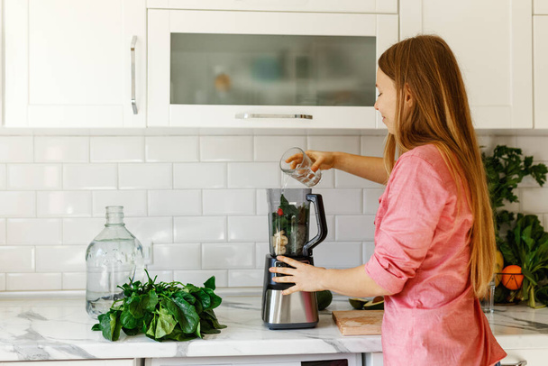 Female smiling, pours water in green smoothie. Healthy food.Happy woman on detox diet, chopped fruits in mixer  - Fotoğraf, Görsel