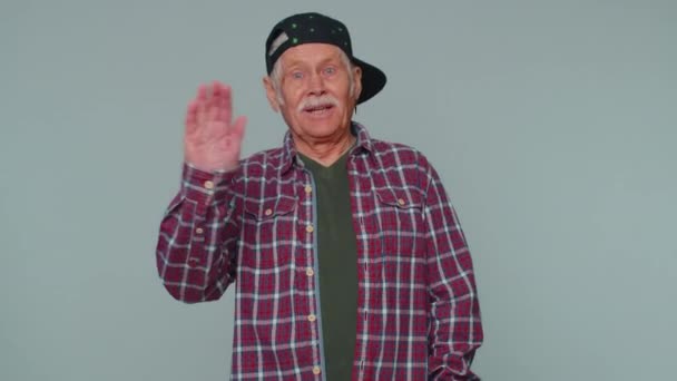 Starszy mężczyzna uśmiecha się przyjazny do kamery i macha rękami gestykulując cześć lub pożegnanie, powitanie - Materiał filmowy, wideo